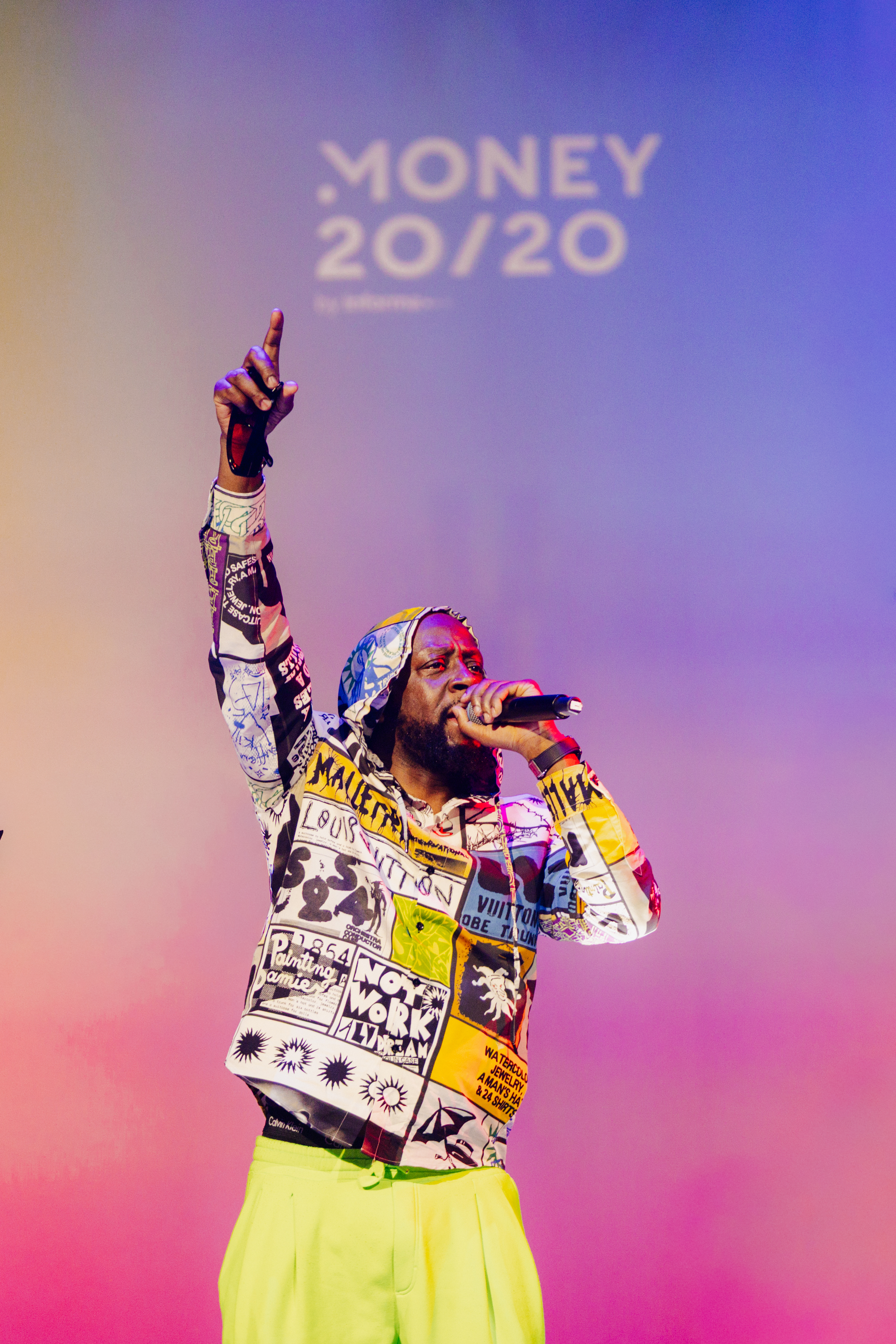 Wyclef Jean performing on stage in a vibrant patterned jacket and neon pants