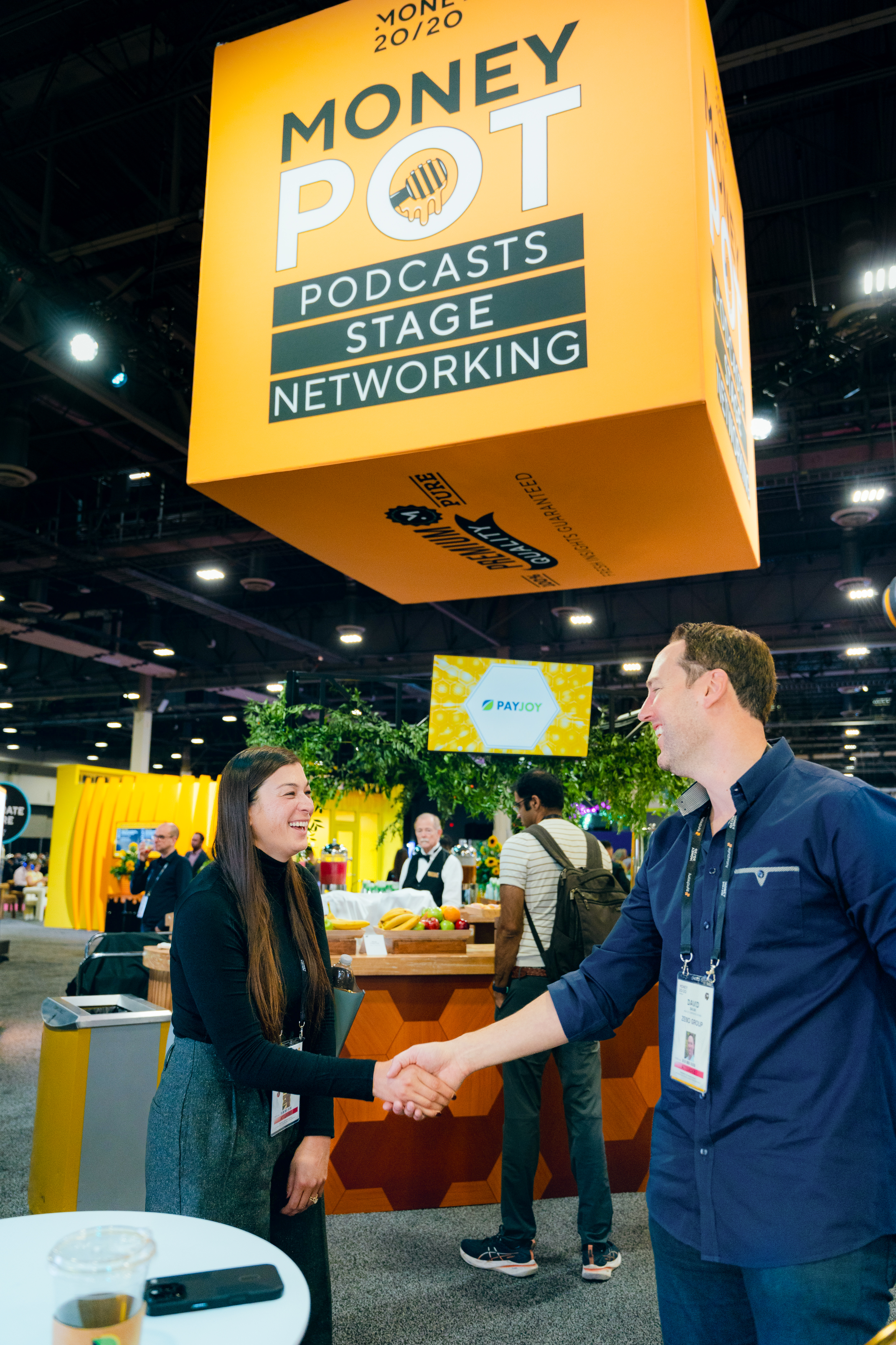 Two attendees shaking hands under the giant yellow Money Pot honey-jar stage signage
